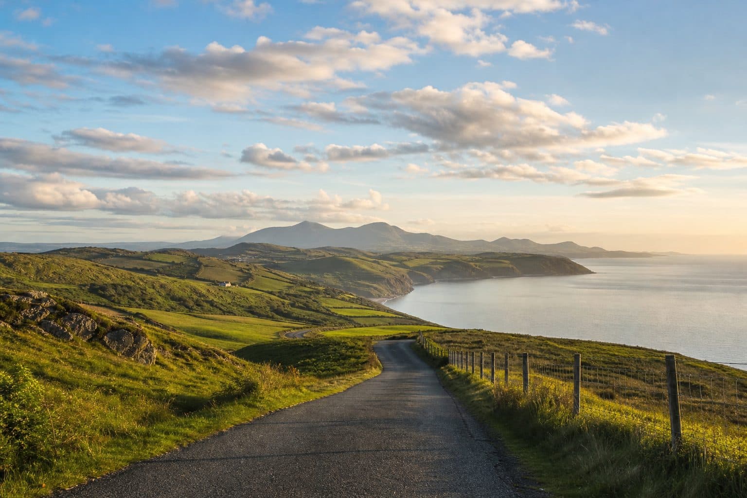 An empty single-track road curving through green hills towards the coast on a bright day, framed for a road trip